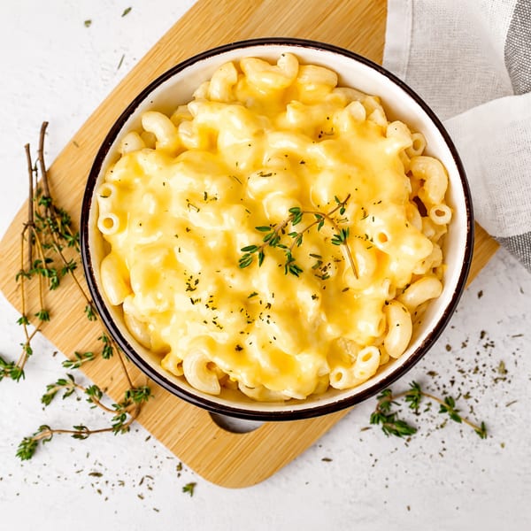 Creamy macaroni and cheese in a white bowl, garnished with fresh thyme, on a wooden board.